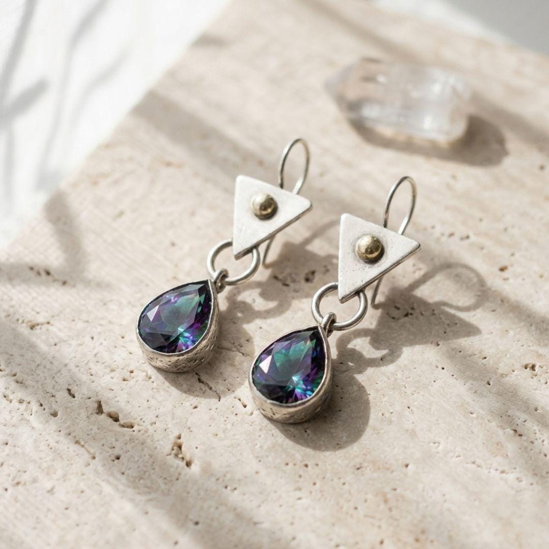 A pair of silver dangle earrings with triangular top charms and teardrop-shaped iridescent gemstones, displayed on a textured beige stone surface with soft shadows.