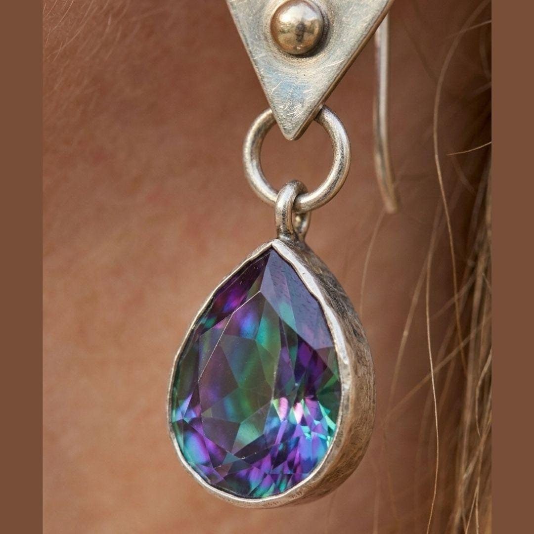 A close-up of a silver-toned teardrop dangle earring with an iridescent faceted gemstone showing purple, green, and blue reflections, worn on a person's ear.