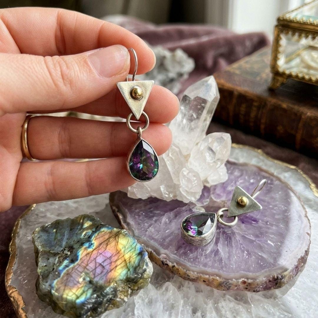 A hand holds a pair of silver geometric dangle earrings with teardrop-shaped iridescent purple gemstones above a crystal cluster and agate slice display.