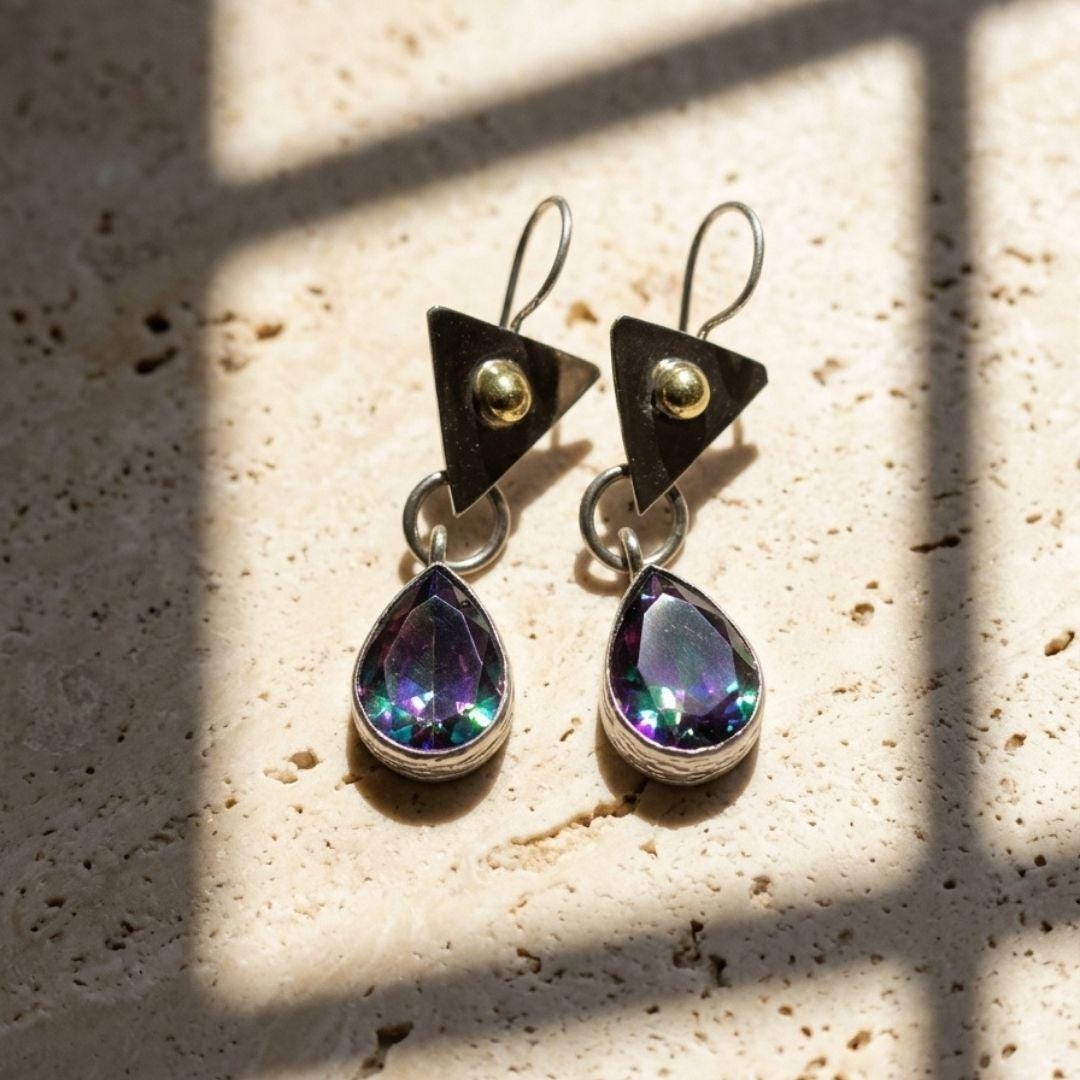 A pair of geometric dangle earrings resting on a textured beige stone surface under warm sunlight with window shadow patterns. The earrings feature triangular black top studs with gold rivets, silver wire hooks, and teardrop-shaped iridescent purple and blue gemstones in silver bezels.